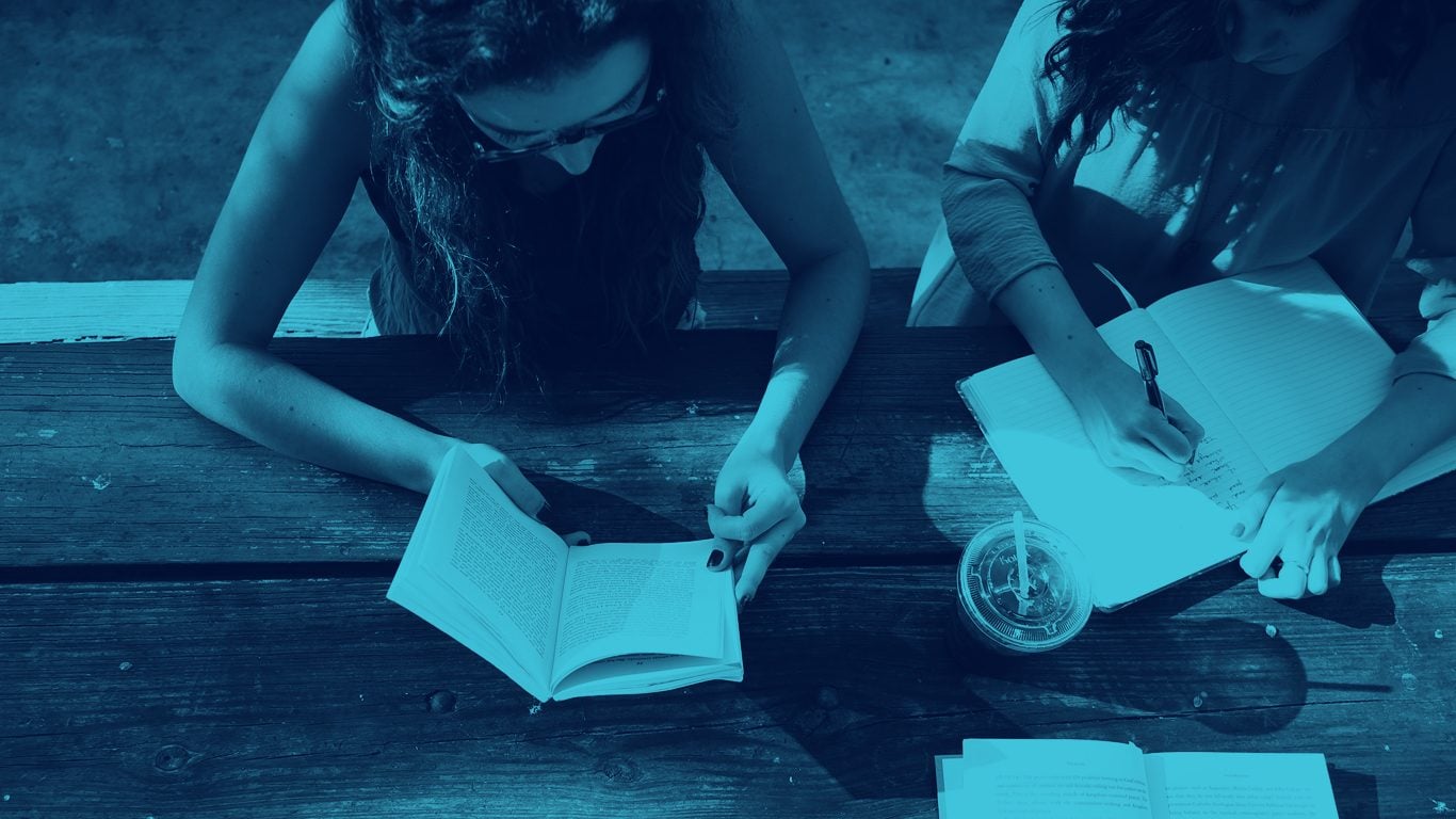 Two women sat at park bench reading and writing