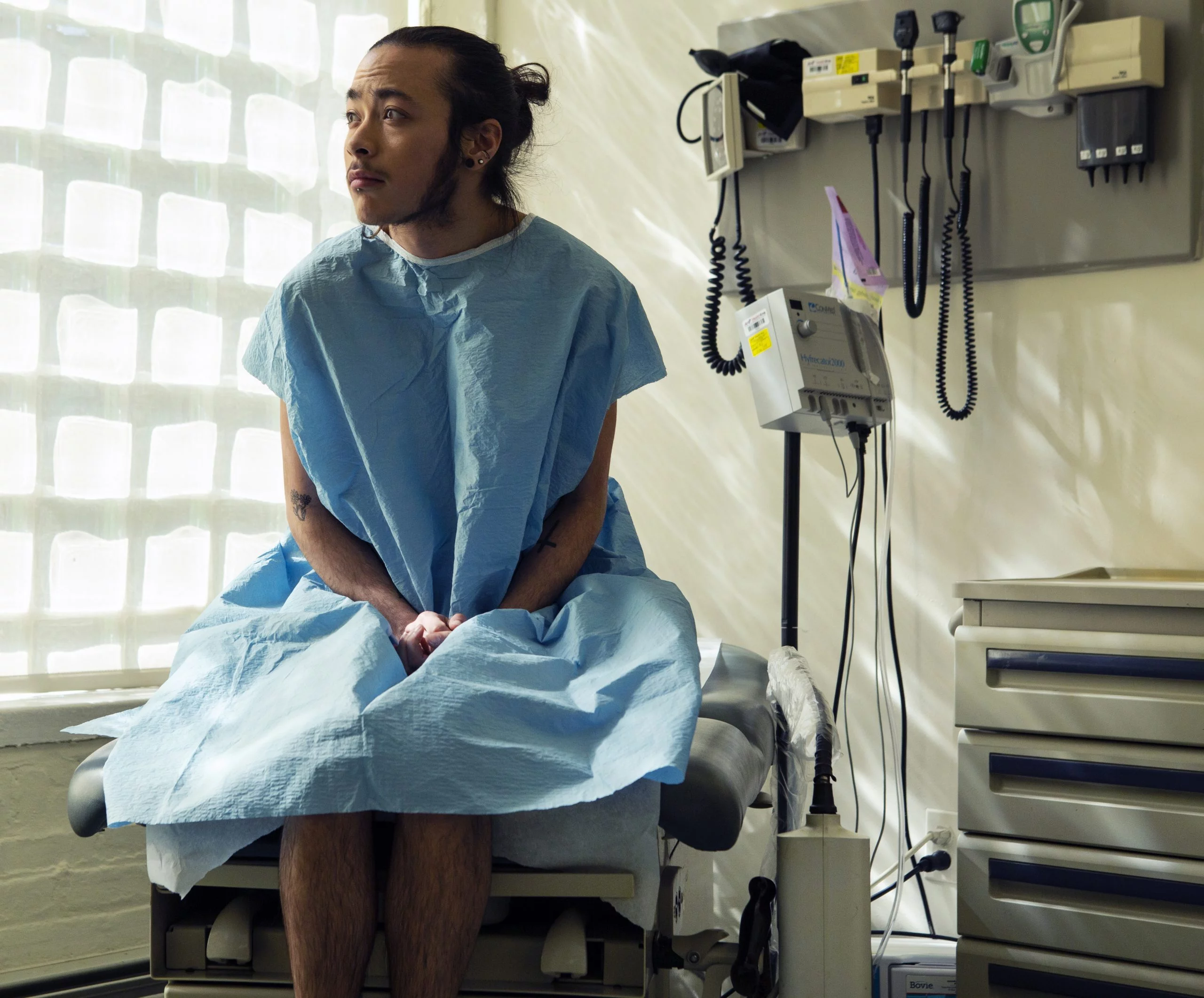 A genderqueer person sitting in a hospital gown