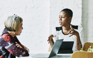 Woman sat at table with laptop while another woman speaks to her