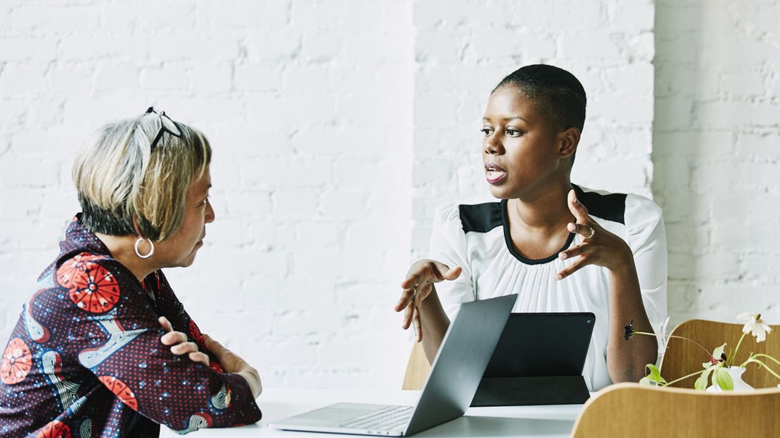 Woman sat at table with laptop while another woman speaks to her