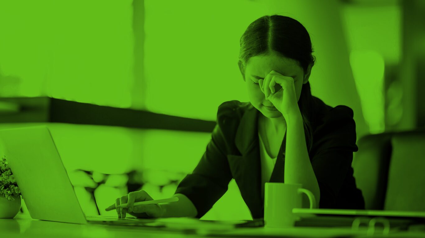 A young woman with black hair wearing a black blazer sits in front of her laptop. Her hand is on her face and she looks stressed.