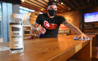 Bartender wiping table with mask on. Bottle of hand sanitizer in front
