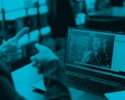 A person sitting at a desk on their laptop communicates with a woman on a Zoom meeting through sign language.