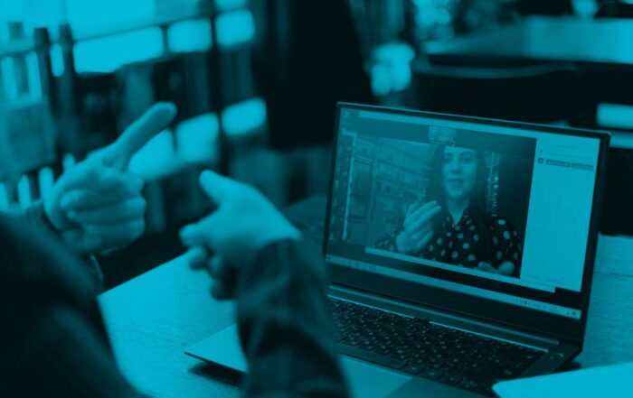 A person sitting at a desk on their laptop communicates with a woman on a Zoom meeting through sign language.