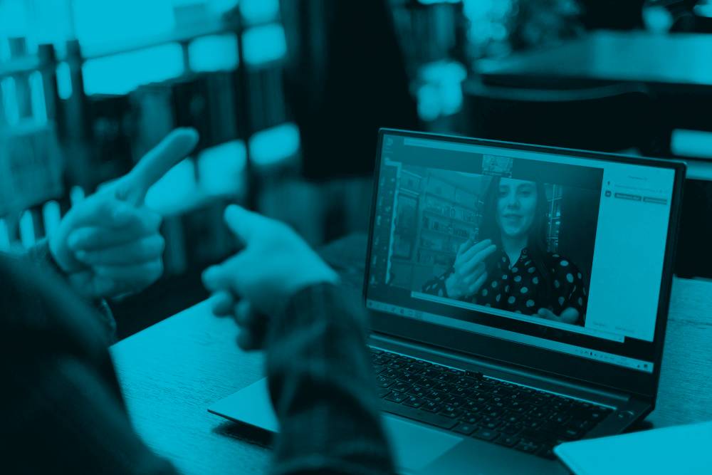 A person sitting at a desk on their laptop communicates with a woman on a Zoom meeting through sign language.