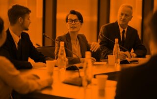 A young woman with short hair and glasses wearing a blazer sits at a business meeting. Two men sit on other side of her listening to her.