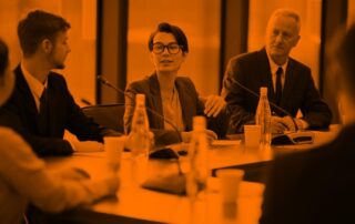 A young woman with short hair and glasses wearing a blazer sits at a business meeting. Two men sit on other side of her listening to her.