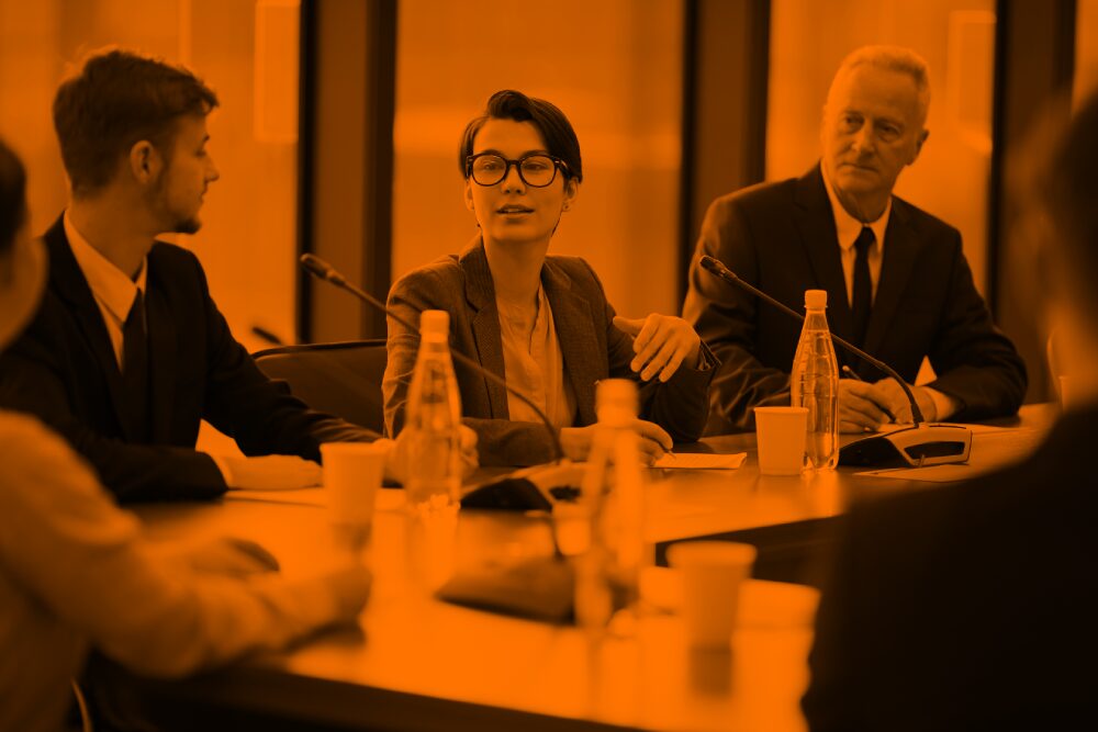 A young woman with short hair and glasses wearing a blazer sits at a business meeting. Two men sit on other side of her listening to her.
