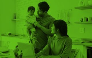 A father holds a baby while a mother works on a laptop