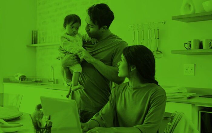 A father holds a baby while a mother works on a laptop