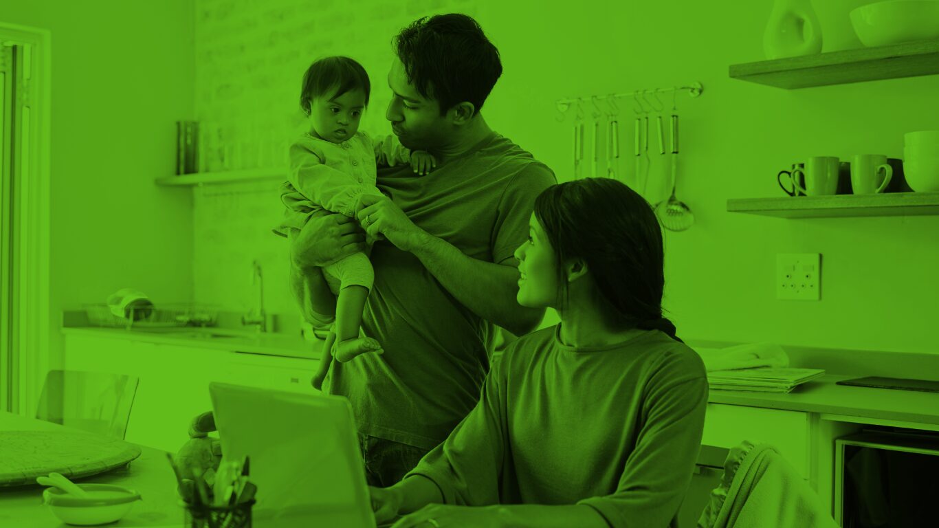 A father holds a baby while a mother works on a laptop