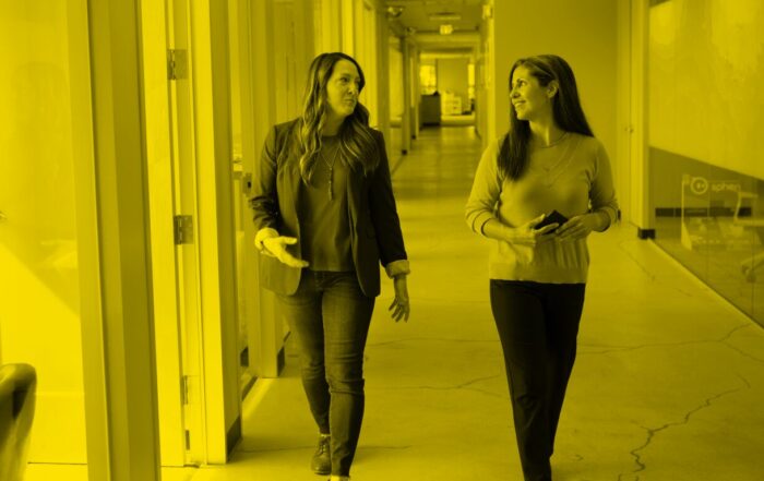Two women walk down a hallway in an office, talking