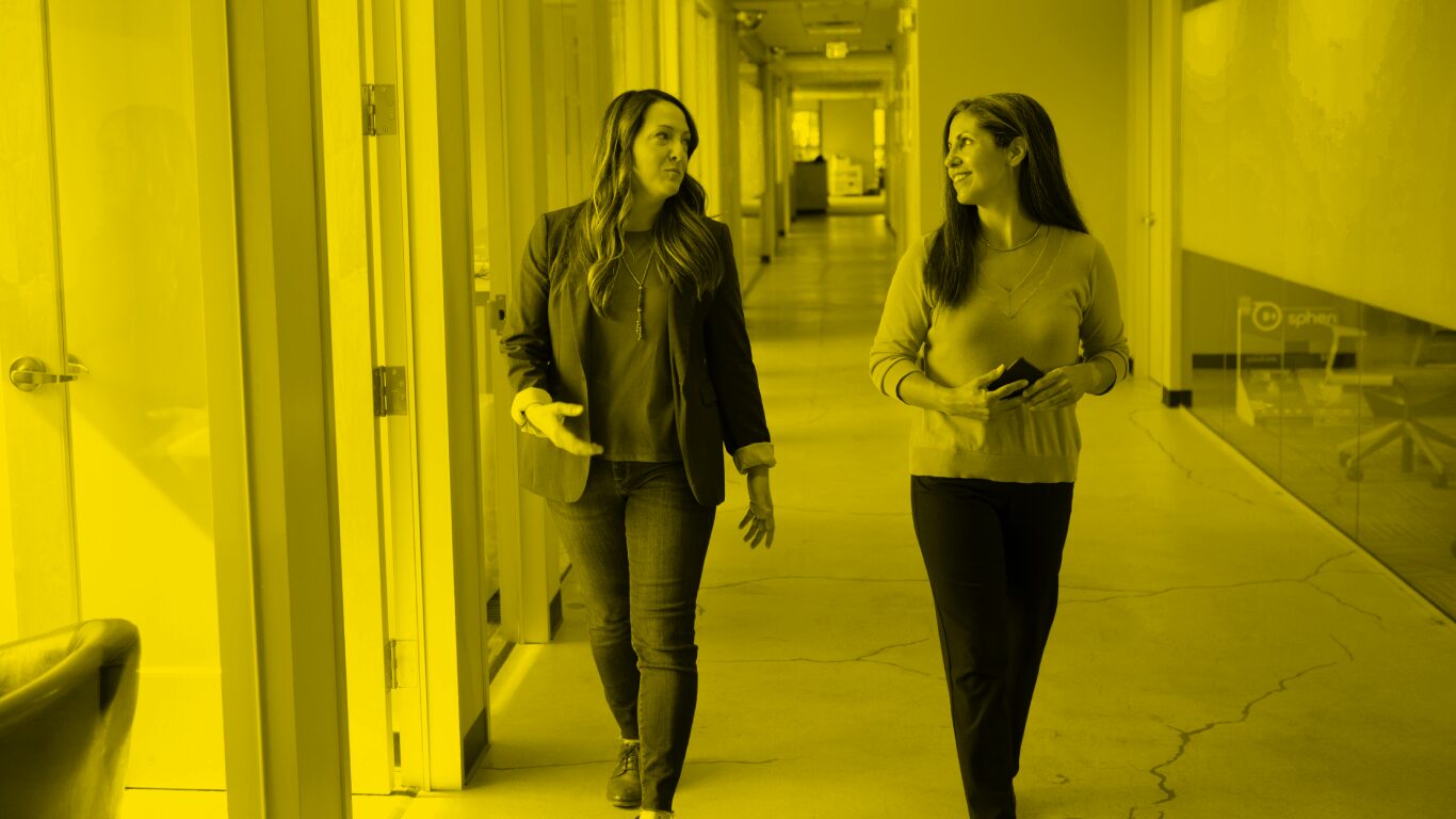Two women walk down a hallway in an office, talking