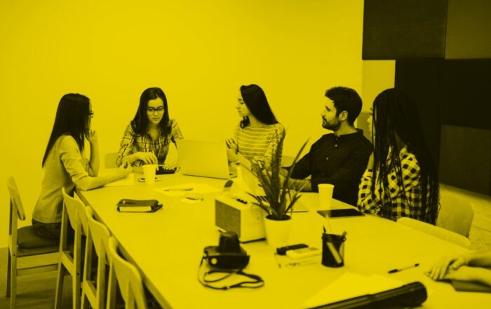 Four employees sit at a table in discussion. A woman wearing glasses is at the head of the table.